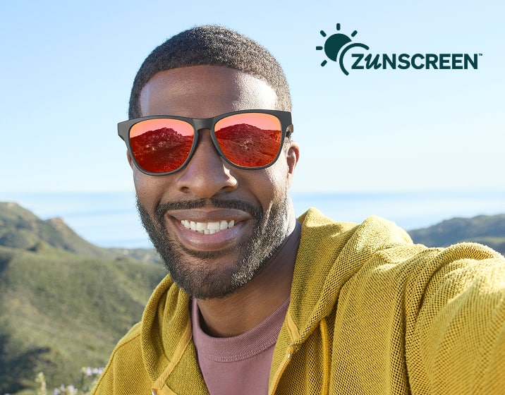 Smiling man taking a selfie outdoors while wearing black wayfarer-style sunglasses with red mirrored lenses, with coastal hills and a blue sky in the background under a "Zunscreen" logo.