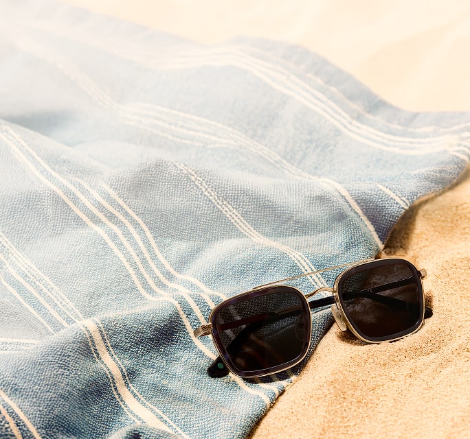 A pair of black square sunglasses with a gold double bridge resting on sand next to a blue and white striped towel.