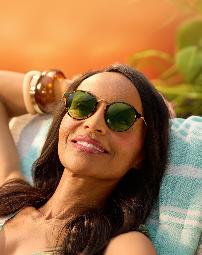 A woman reclining on a striped beach towel wearing round tortoiseshell sunglasses with green-tinted lenses and a gold nose bridge.