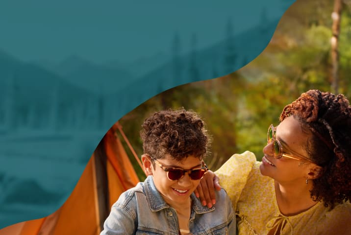 A smiling woman in translucent amber sunglasses and a young boy in tortoiseshell-patterned shades with dark red lenses, posed in front of a golden-hour outdoor scene.