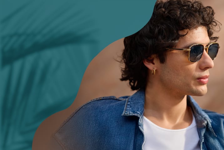 A man in a denim jacket wearing square gold-framed aviator sunglasses with dark gradient lenses, featuring a textured metallic double bridge.