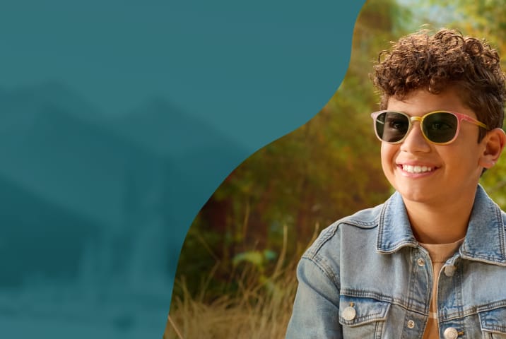 A young boy smiling outdoors wearing round-frame sunglasses with a pink-to-yellow gradient frame and dark protective lenses.