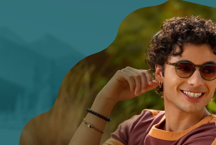 A man with curly hair wearing round olive-green acetate sunglasses with brown gradient lenses, paired with a maroon and orange t-shirt.