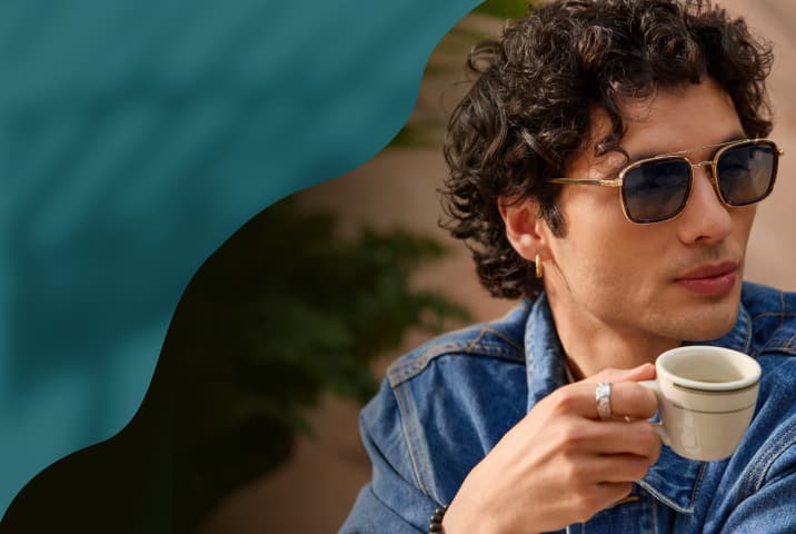 A close-up of a man wearing rectangular gold-framed sunglasses with dark lenses and a double bridge, holding a white coffee cup.