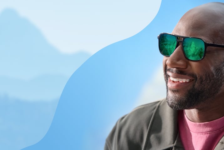 A man wearing black-framed aviator sunglasses with vibrant green mirrored polarized lenses, reflecting a mountain landscape.