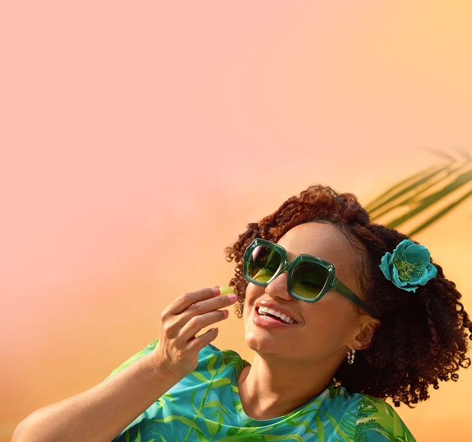 Smiling woman with curly hair and a teal flower accessory wearing oversized green geometric sunglasses against an orange and pink gradient background.