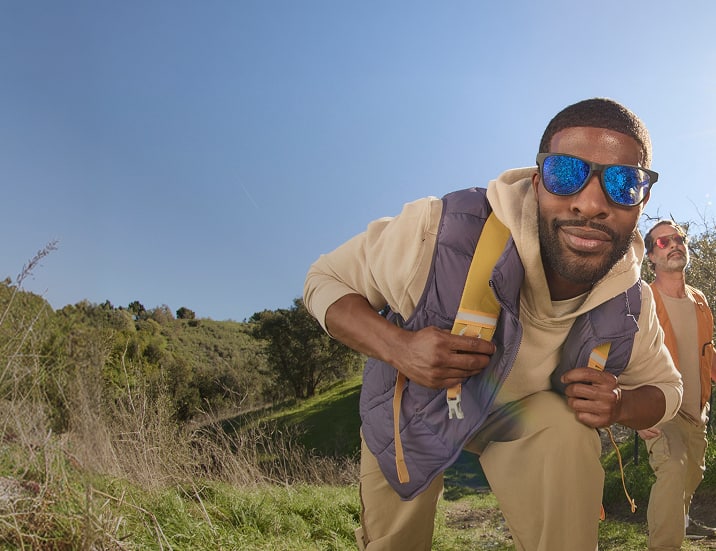A man in hiking clothes wearing black square sports sunglasses with blue mirror frames. Another man is in the background wearing tortoiseshell aviator sports sunglasses with rose gold mirror lenses. Background is green rolling hills.