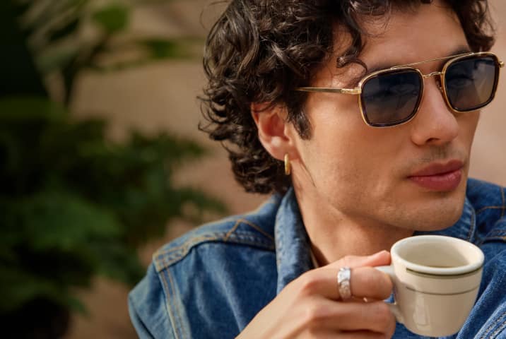 A man in gold square aviator Zenni sunglasses. He has mid-length curly brown hair and is sipping an espresso.