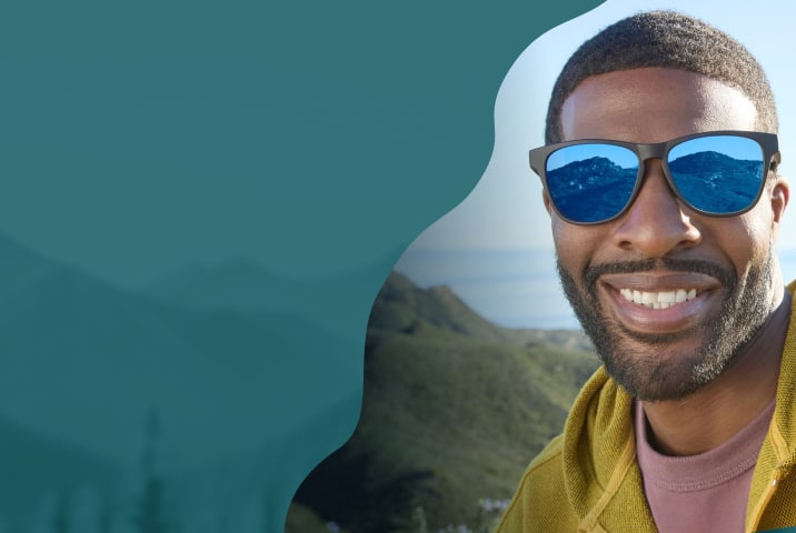 A man with close cropped hair and a beard wearing square sports sunglasses with blue mirror lenses. Background is rolling hills and the ocean.