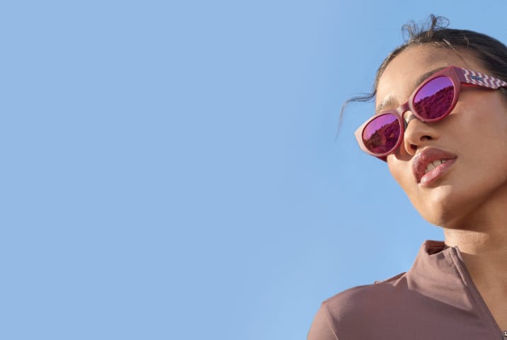 A woman wearing pink oval sports sunglasses with patterned temples and pink mirror lenses. Blue sky background.