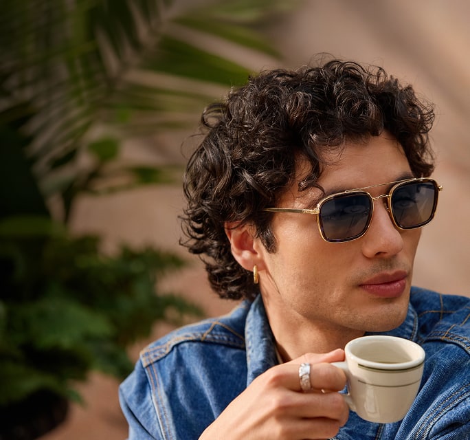 A man in gold square aviator Zenni sunglasses. He has mid-length curly brown hair and is sipping an espresso.