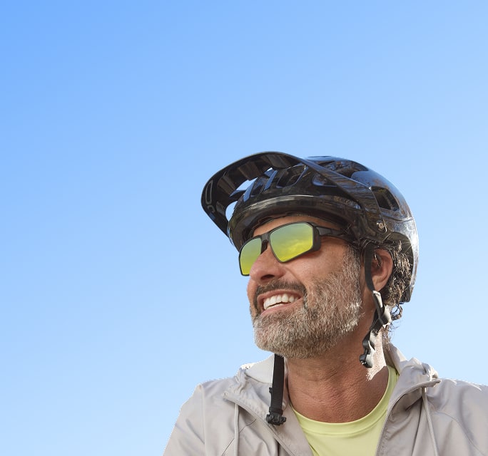 A smiling man with a grey beard wearing a black cycling helmet and matte black sport sunglasses with yellow mirrored lenses against a clear blue sky.
