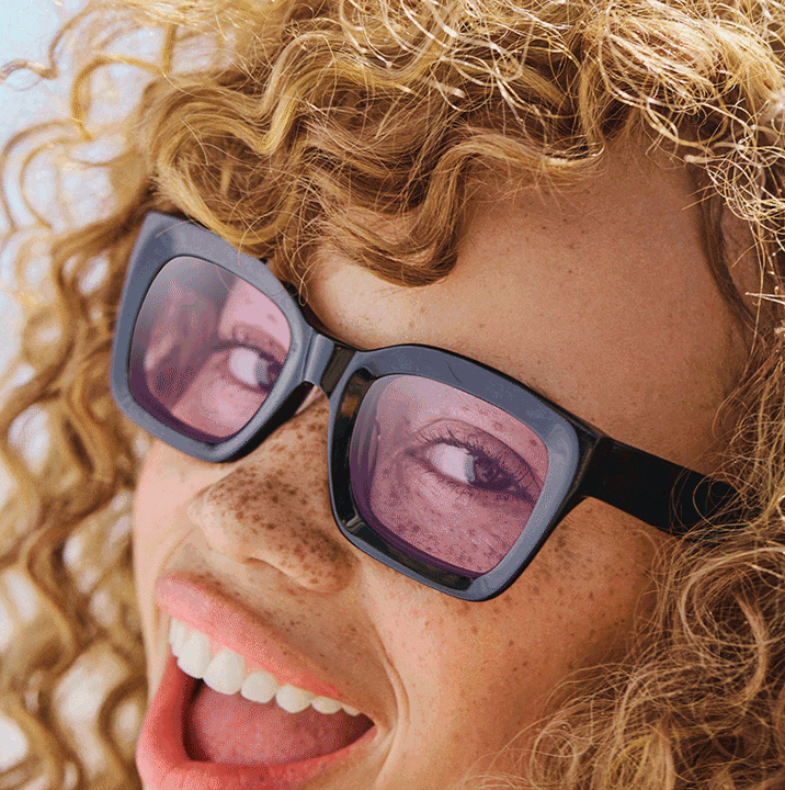 A woman with curly hair and freckles wearing black thick rim square sunglasses, with lenses cycling through various tints to show options.