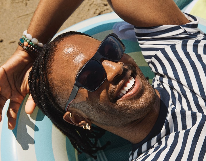 A man with box braids wearing black square sunglasses with dark gray lenses, reclining on a pool floatie at the beach.