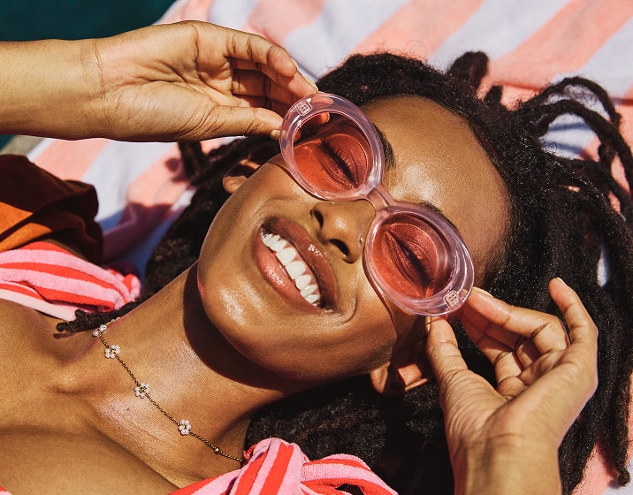 A woman with locs wearing oversized pink oval pink sunglasses with pink lenses, relaxing on a pink and white striped beach towel next to a pool.