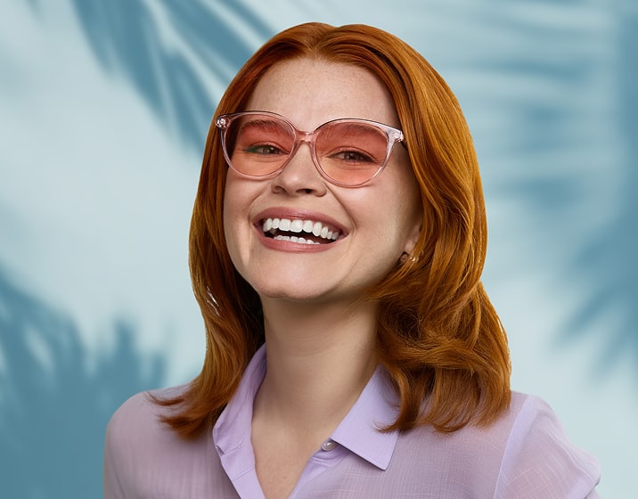 A woman with long red hair wearing pink round plastic glasses frames with pink lenses, standing in front of a wall with shadows of a palm tree.