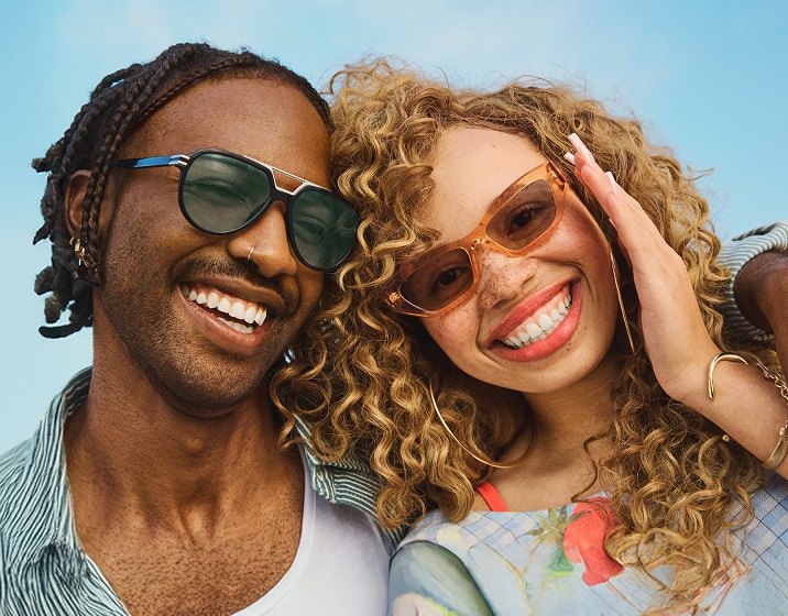 A man with box braids wearing blue and black aviator sunglasses and a woman with curly blond hair wearing orange oval sunglasses.