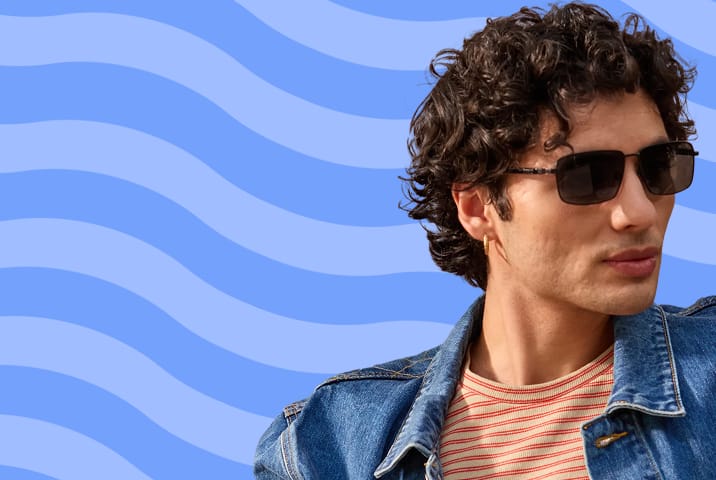 Profile of a man with wavy dark hair wearing rectangular metal-framed sunglasses and a denim jacket, set against a light blue background with abstract darker blue waves.
