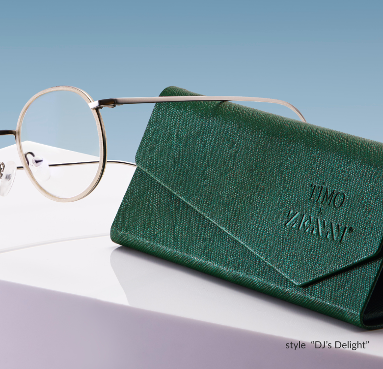 A pair of Timo x Zenni round metal glasses in style “DJs Delight” placed next to a green eyeglass case against a blue background.