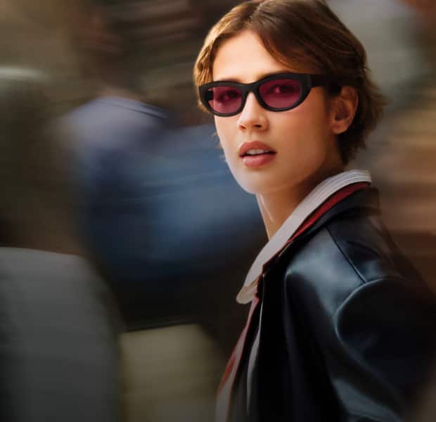 Woman with short brown hair wearing narrow black sunglasses with pink-tinted lenses. They are dressed in a black leather jacket over a collared shirt and are looking slightly to the side. The blurred background suggests a busy urban scene, giving the photo a sense of motion and energy.