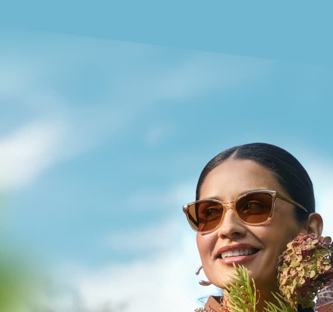 A smiling woman with dark hair pulled back holding a large bouquet of pink and purple flowers while wearing translucent brown cat-eye glasses with amber-tinted lenses against a soft-focus blue sky background.