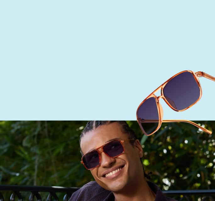 A smiling man with dark hair in small braids wearing translucent orange aviator glasses with deep amethyst-tinted lenses, positioned against a background of lush green foliage and a clear blue sky.