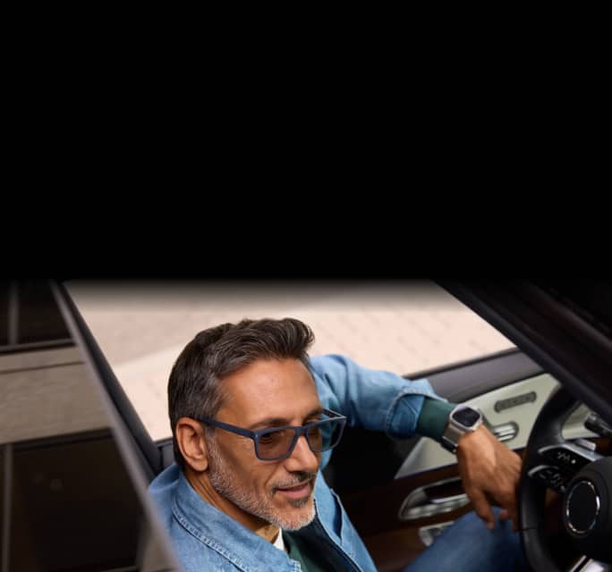 High-angle shot through a car's sunroof of a middle-aged man with salt-and-pepper hair sitting in the driver's seat, wearing a denim shirt and dark grey rectangular eyeglasses with lightly tinted transition lenses.