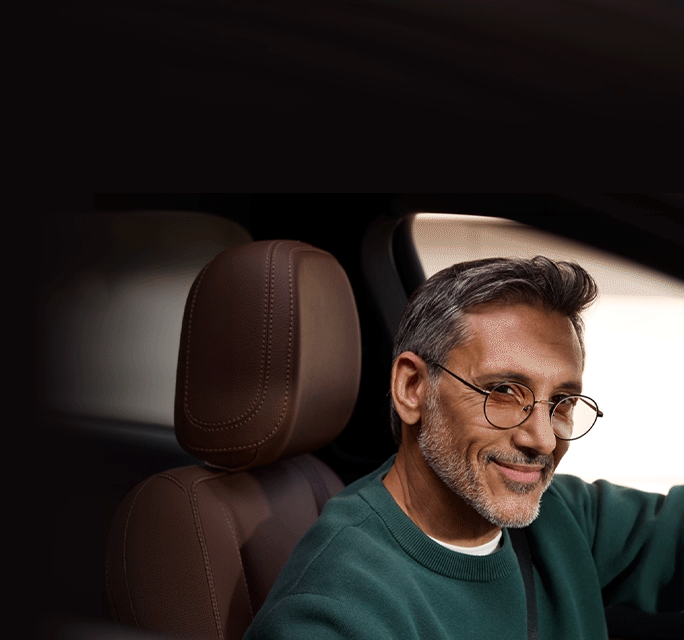 Side-angle view of a smiling middle-aged man with salt-and-pepper hair and a beard sitting in the driver's seat of a car, wearing round wireframe eyeglasses and a green sweater.