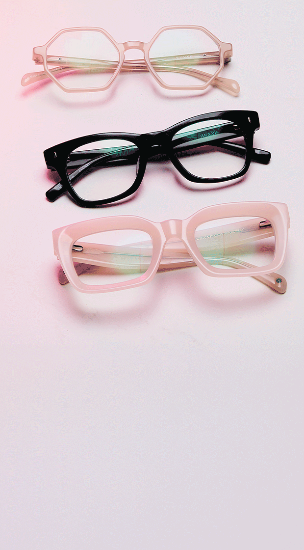 Three pairs of stylish eyeglasses—octagonal blush pink, classic black rectangular, and bold square light pink—are neatly arranged on a soft pink gradient surface with a subtle reflection.