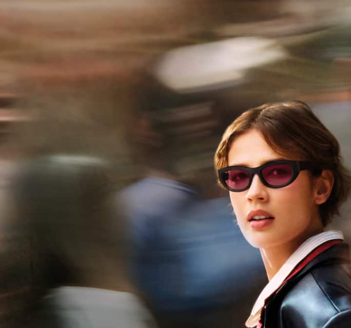 Woman with short brown hair wearing narrow black sunglasses with pink-tinted lenses. They are dressed in a black leather jacket over a collared shirt and are looking slightly to the side. The blurred background suggests a busy urban scene, giving the photo a sense of motion and energy. A graphic displays a lens transitioning through clear to dark red shades, illustrating adaptive lens technology. The text reads 'Ultra Dynamic' and 'Light Intelligent Lenses', emphasizing their responsive design.