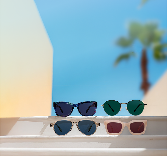 Four pairs of sunglasses on a white ledge with a gradient sky and a blurred palm tree in the background.