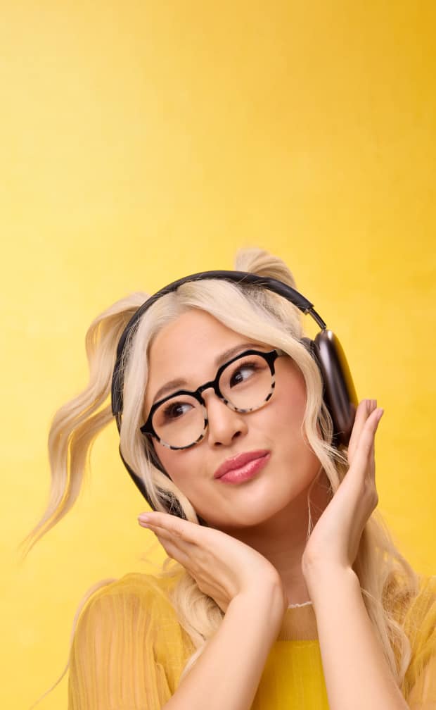 A woman resting her chin on her hand while wearing delicate rimless glasses with a gold bridge and arms against a wavy white background.
