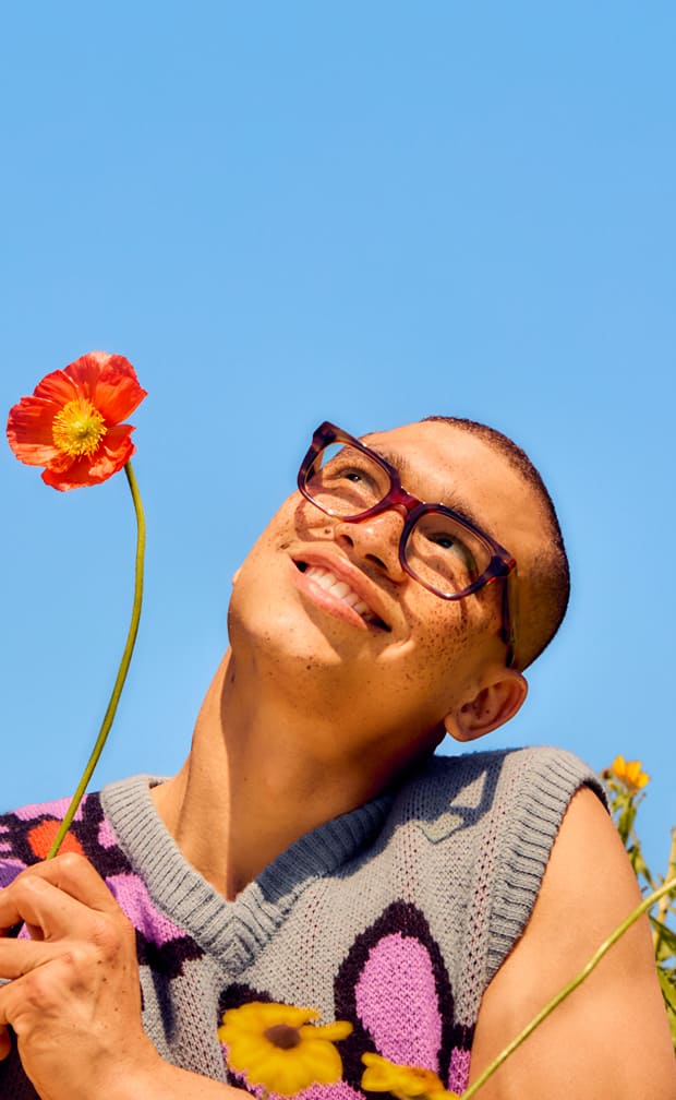 A smiling woman with long dark hair and freckles wearing oversized, translucent pink cat-eye glasses against a light blue sunburst background.