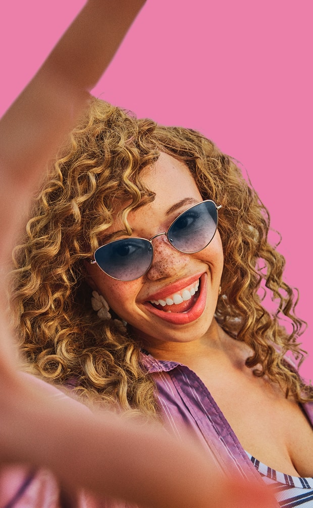 A woman with blonde curly hair smiling and reaching toward the camera while wearing gold cat-eye sunglasses with blue-tinted lenses against a pink background.