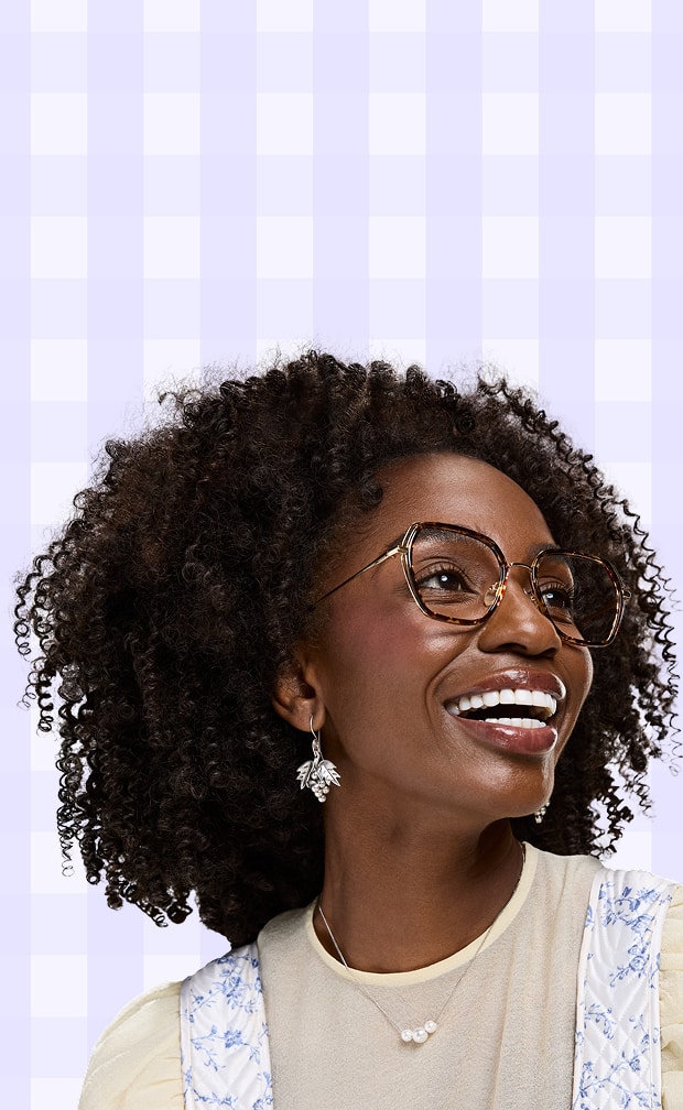 A laughing woman with dark curly hair looking upward while wearing thin gold geometric glasses against a light purple checkered background.