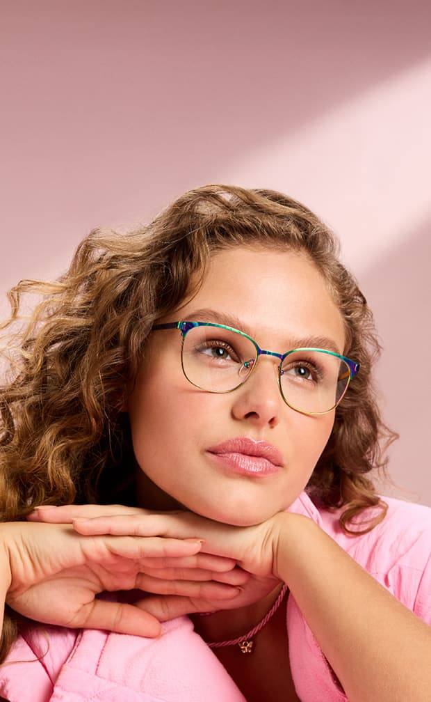 A woman with blond curly hair wearing rainbow metal square Zenni glasses, on a pink background.