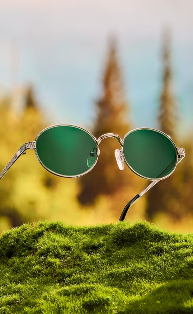 A pair of gold oval glasses with green-tinted lenses standing upright in green moss against a blurred background of evergreen trees.