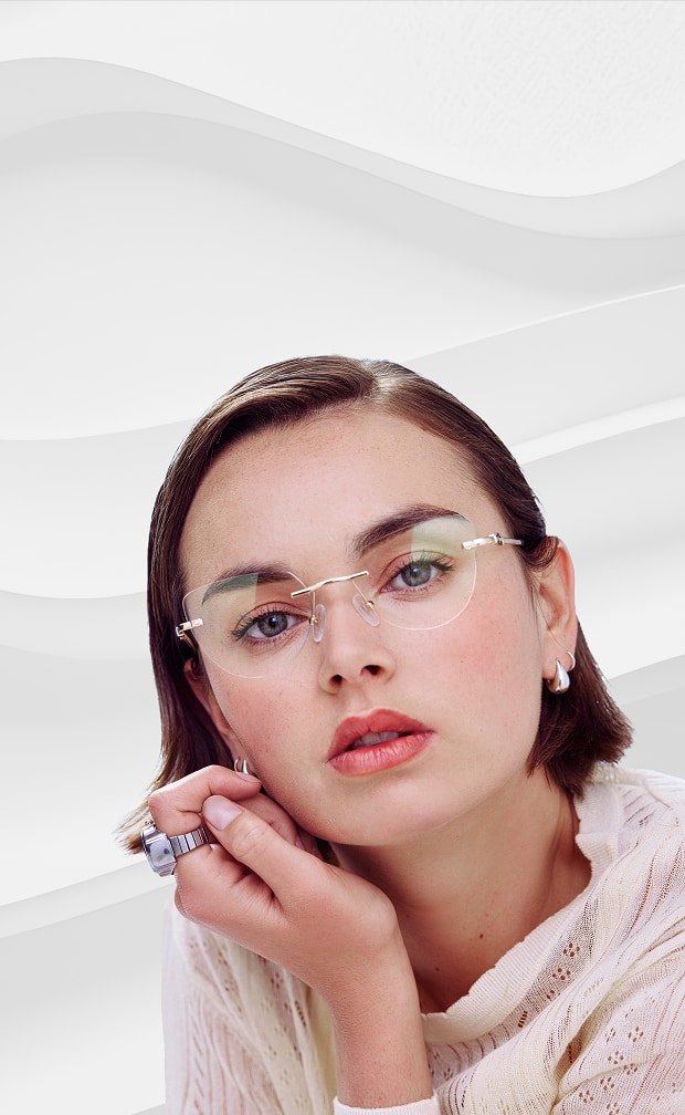 A woman resting her chin on her hand while wearing delicate rimless glasses with a gold bridge and arms against a wavy white background.
