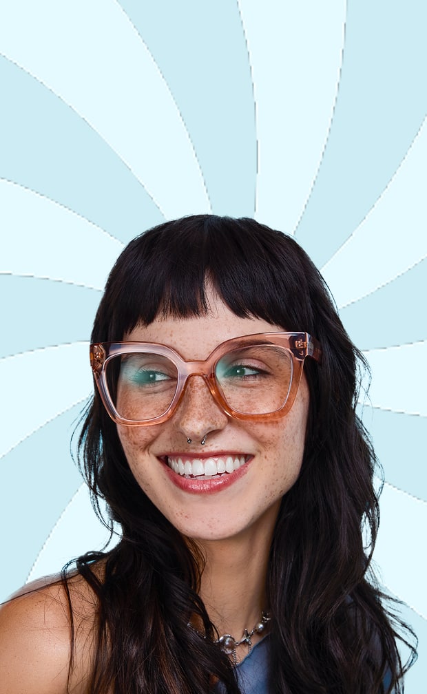 A smiling woman with long dark hair and freckles wearing oversized, translucent pink cat-eye glasses against a light blue sunburst background.