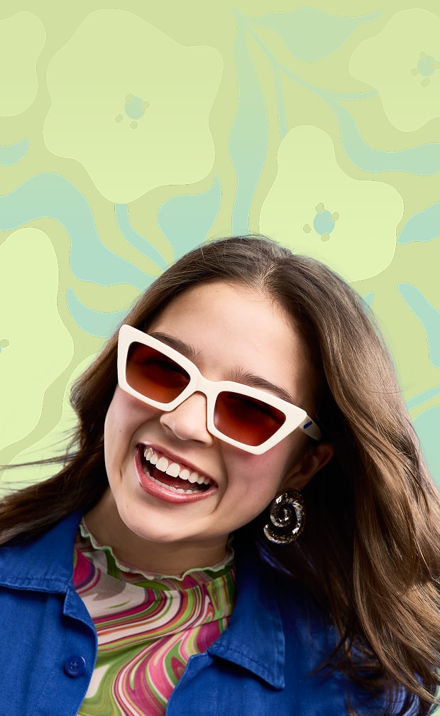 A smiling woman with long dark hair and freckles wearing oversized, translucent pink cat-eye glasses against a light blue sunburst background.