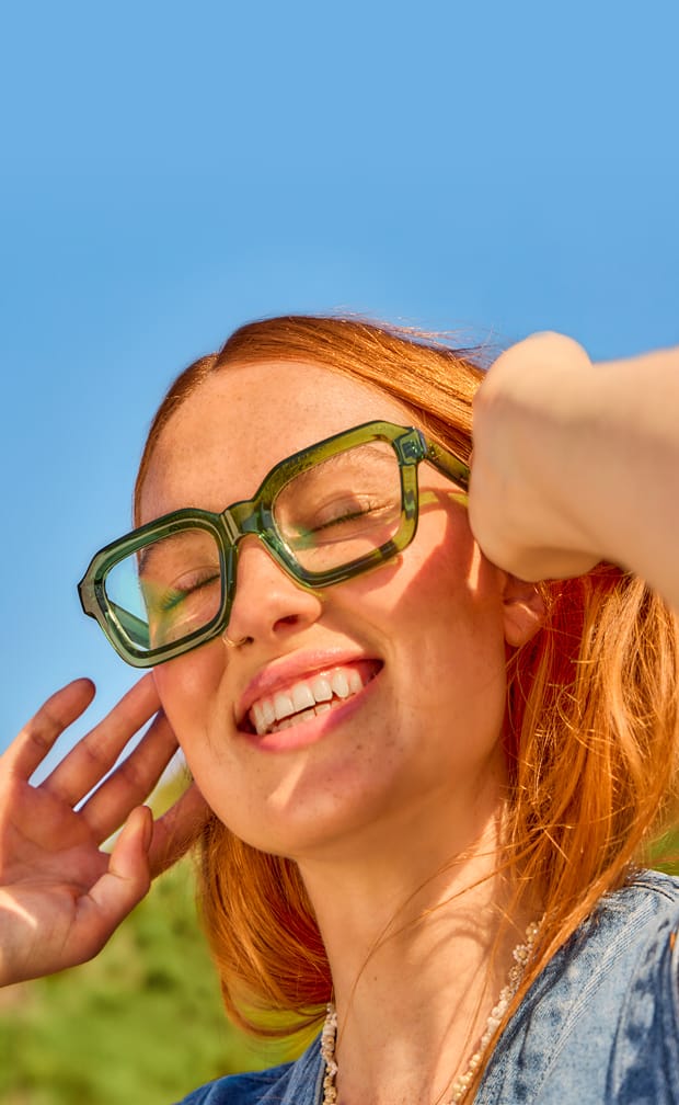 Red-haired woman in oversized Zenni green glasses, smiling under bright sunlight.
