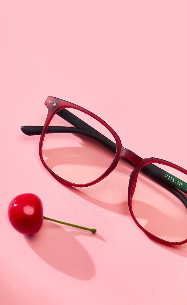 Red round Zenni glasses next to a cherry on a pink background.