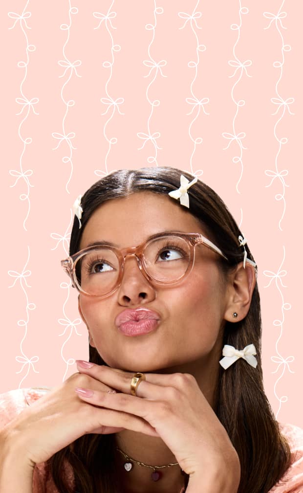 Woman in Zenni pink translucent glasses with white hair bows, posing playfully on a pink background.