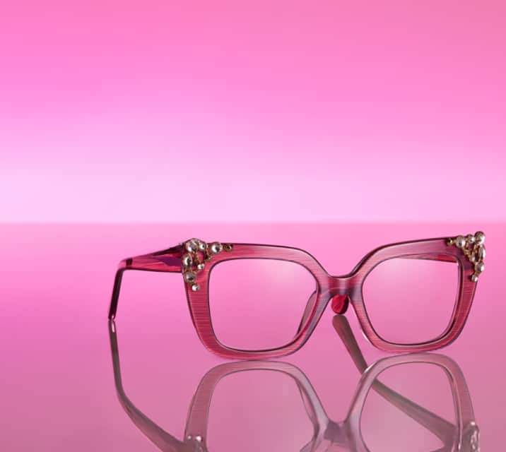  A pair of oversized pink Zenni glasses with temple embelleshments on a pink reflective surface with a soft glow.