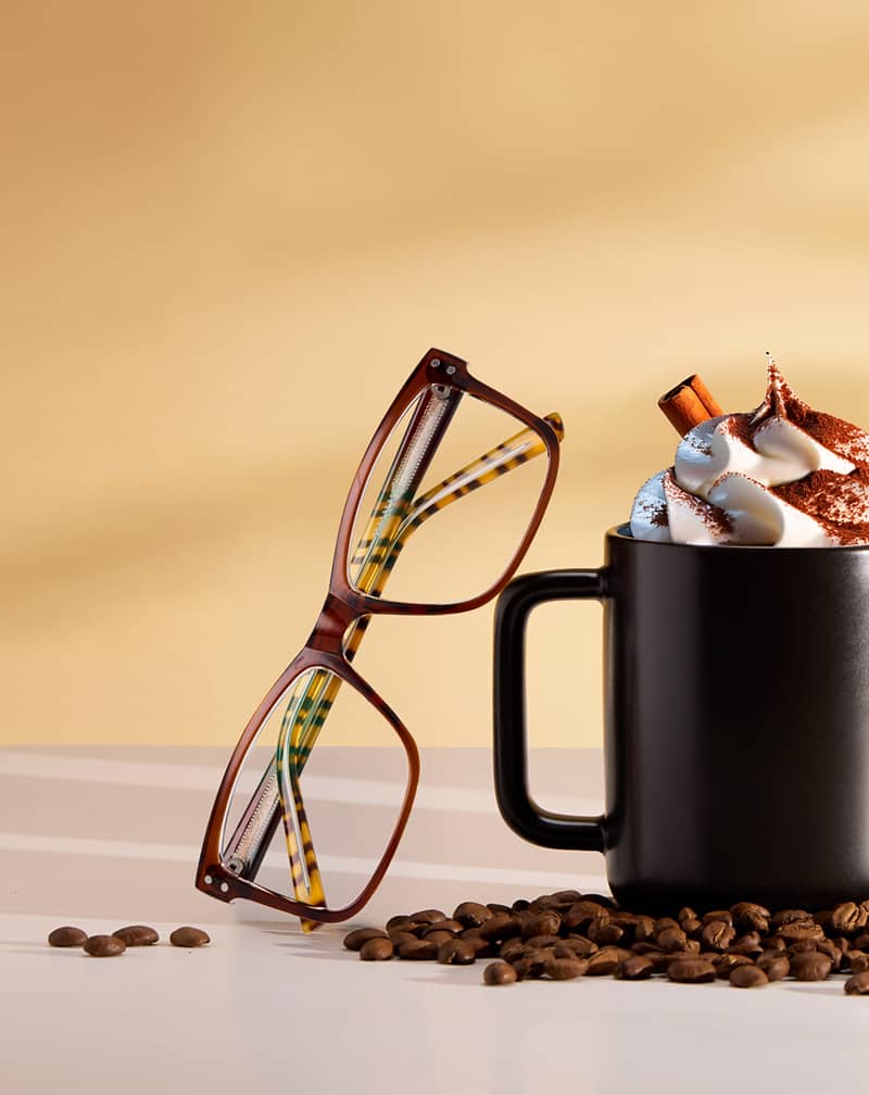 Tortoiseshell Zenni glasses leaning against a black mug of whipped coffee topped with cinnamon on a beige background.