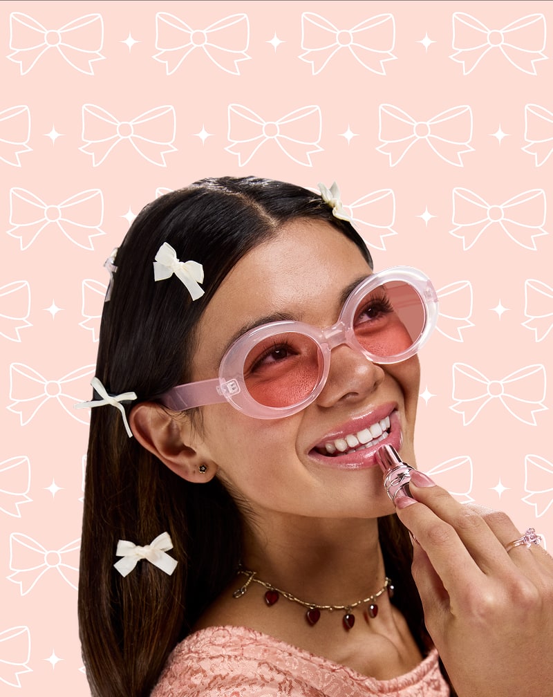 Woman in Zenni pink translucent glasses with white hair bows, posing playfully on a pink background with illustrated bows.
