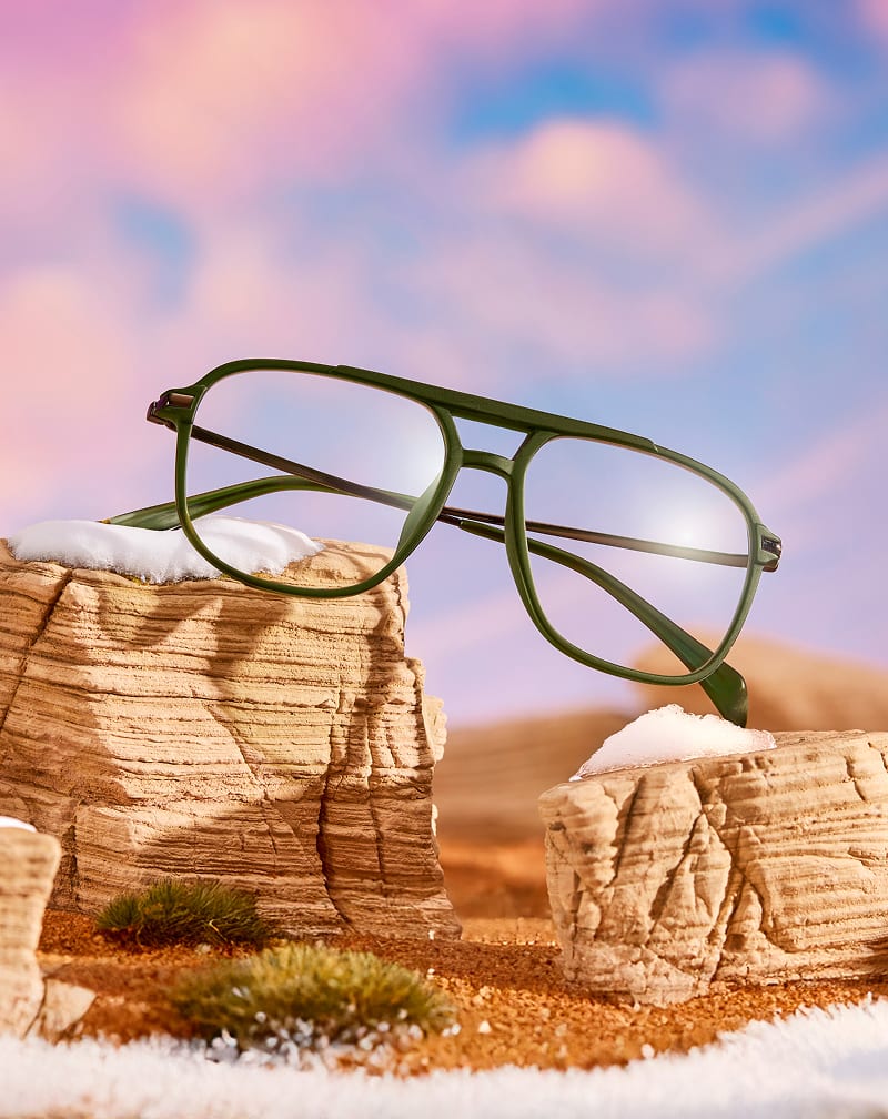Zenni green aviator-style eyeglasses resting on desert rocks dusted with snow under a pastel sky.