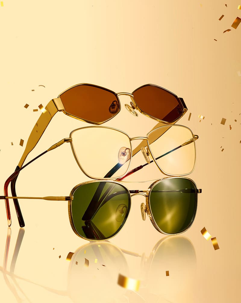 Three pairs of Zenni gold-rimmed eyeglasses and sunglasses stacked together amid falling gold confetti on a warm golden backdrop.