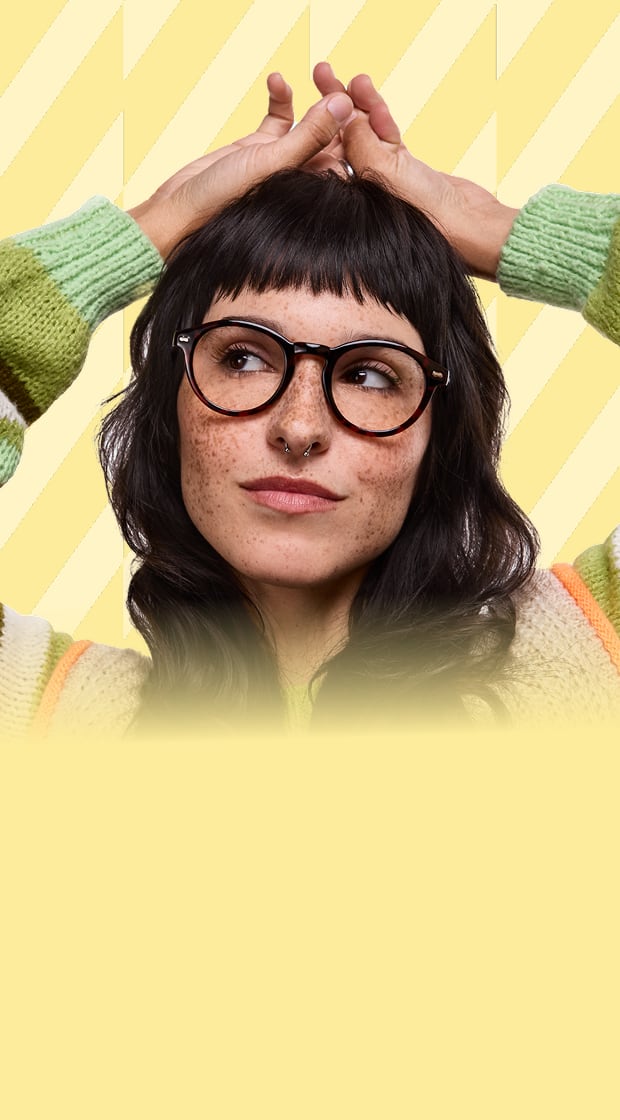 The image features a woman with dark wavy hair and freckles wearing round glasses with sleek, glossy black frames. She is dressed in a cozy, multicolored striped sweater with shades of green, white, and brown. Her hands rested on her head.