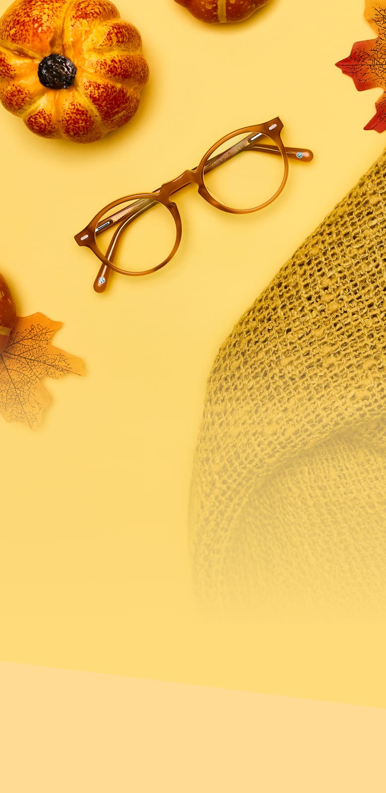 Round brownish orange glasses on a yellow surface next to an orange pumpkin, an orange leaf, and the corner of a burlap table cloth.